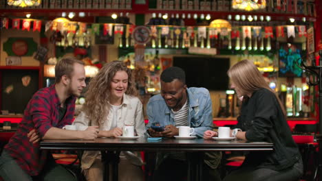 Retrato-De-Jóvenes-Amigos-Alegres-Mirando-Un-Teléfono-Inteligente-Mientras-Están-Sentados-En-Un-Café.-Personas-De-Raza-Mixta-Sentadas-En-Una-Mesa-En-Un-Restaurante-Usando-Un-Teléfono-Móvil.